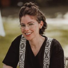 A person with brown hair and glasses wearing a black shirt and a black and white vest is smiling.